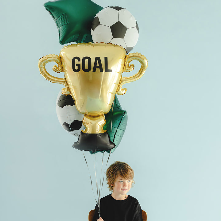Junge sitzt unter dem schwebenden Fußball-Ballonstrauß mit goldenem GOAL-Pokal, Fußbällen und grünen Sternen